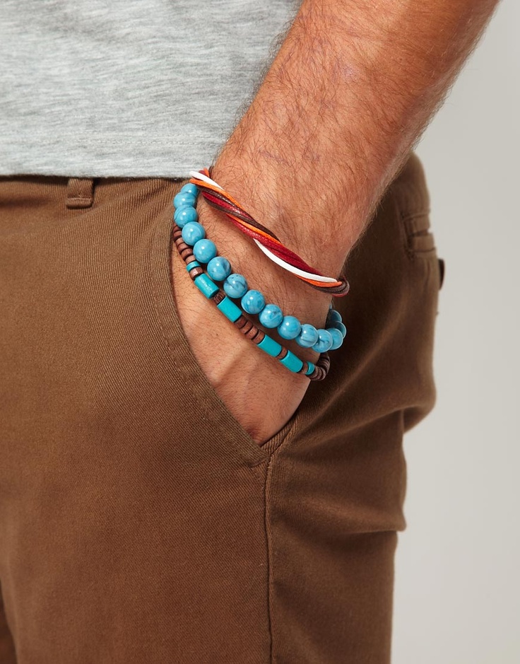 a closeup shot of a man's wrist wearing three bracelets. the top bracelet is a white and red leather cord bracelet. the middle bracelet is a blue beaded bracelet with a wooden accent. the bottom bracelet is a brown leather bracelet with a silver accent. the man is wearing brown pants. the background is a blurry white.