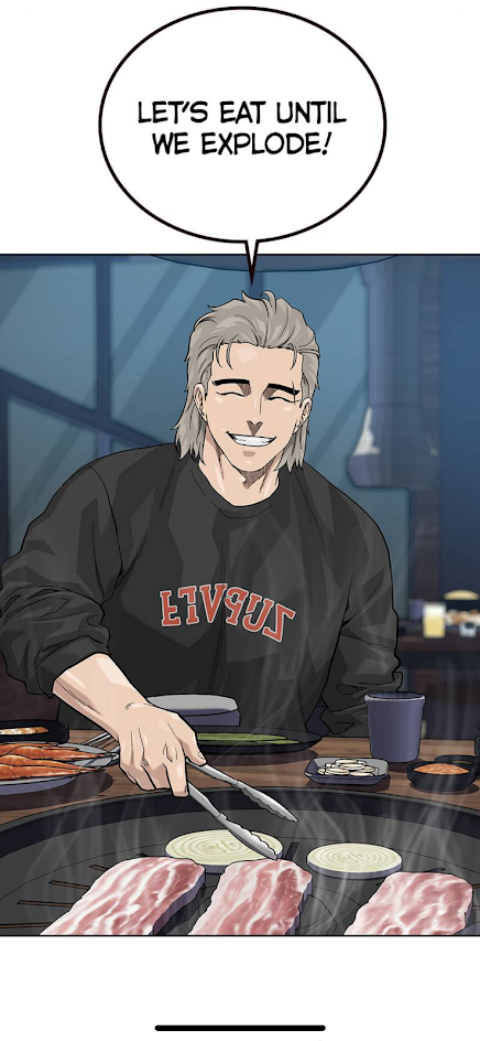 a man wearing a black sweatshirt with a white logo that's flipped is smiling widely while holding a pair of tongs. there is a hot grill with meat and onions on it. the man is sitting in a restaurant setting. there is a speech bubble above his head that says "let's eat until we explode". he appears to be enjoying the meal.