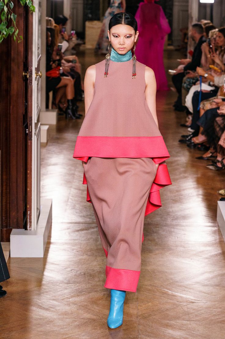 a female model walks the runway wearing a pink and blue dress, with pink trim and matching boots. the dress is sleeveless and has a gathered skirt. she is also wearing large tassel earrings. the model is looking straight ahead as she walks.