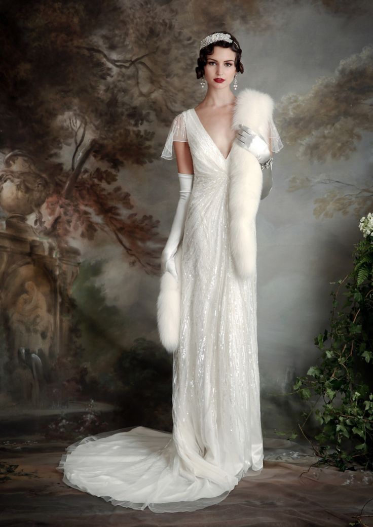 a woman in a white wedding dress with a fur stole stands in front of a painted background. she is wearing white gloves and has her hair styled in a retro updo. the dress has a vneckline and a flowing train. the painting on the wall depicts a landscape with trees and flowers. the woman's red lipstick and glamorous look are reminiscent of the art deco era.
