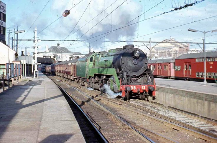 a vintage photograph depicting a green steam locomotive pulling a passenger train through a railway station, possibly in cape town, south africa.  the train is composed of several carriages, some of which are visible behind the engine.  the station platform is visible, along with overhead power lines and other railway infrastructure.  there are buildings in the background, and the overall scene suggests a bustling railway environment of a bygone era.  the image has a slightly faded, vintage quality.