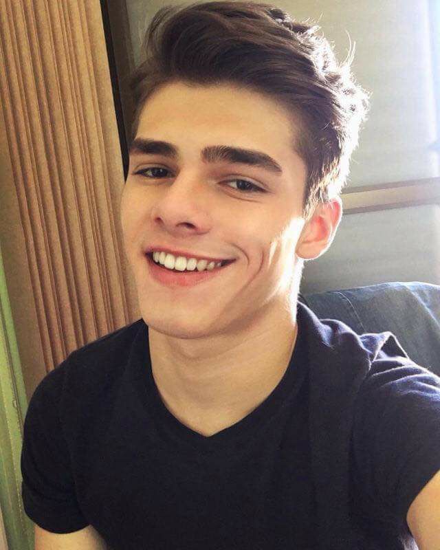 a young man with brown hair is wearing a black tshirt, he is looking at the camera and smiling. the image is a close up and taken indoors.