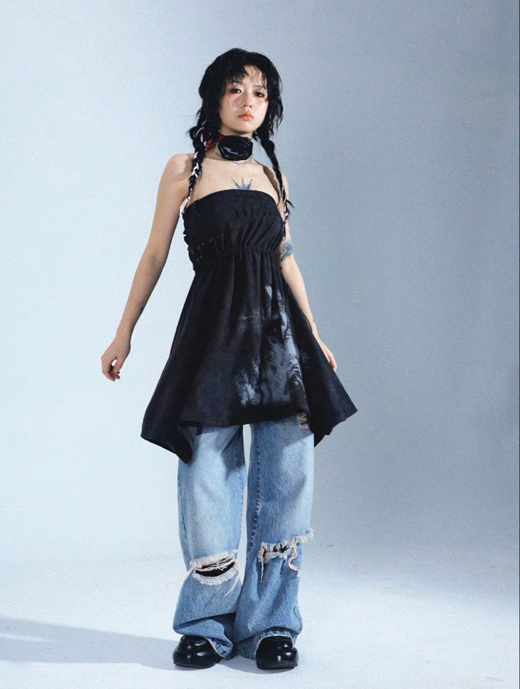 a fullbody shot of a young woman posing in a studio setting. she's wearing a black asymmetrical strapless dress with a subtle tiedye pattern, paired with light blue wideleg jeans that feature distressed details at the knees and hems.  she's also sporting a black choker necklace and her dark hair is styled with loose braids. the background is a neutral light gray. the overall style is edgy and fashionable, blending grunge elements with contemporary trends.