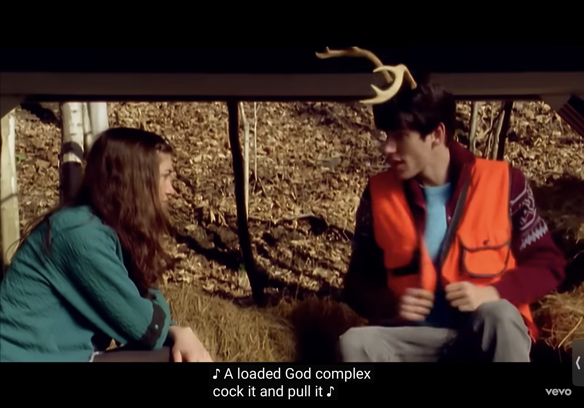 a man and a woman are sitting on a pile of hay in a forest. the man is wearing an orange hunting vest and has a deer antler on his head. the woman is looking at him and he is talking to her and laughing.  the woman is looking down and is wearing a teal shirt.  the forest is full of fall leaves and the hay looks like it could be from a hay bale.   the man is looking like he is about to tell a joke or a story.