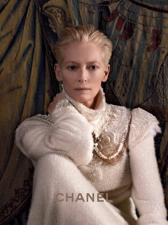 a blonde woman is wearing a white sweater with a fur collar and pearls, she looks at the camera with a serious expression. the background is out of focus and features a tapestry. the image is taken close up, showcasing the details of the woman's outfit.