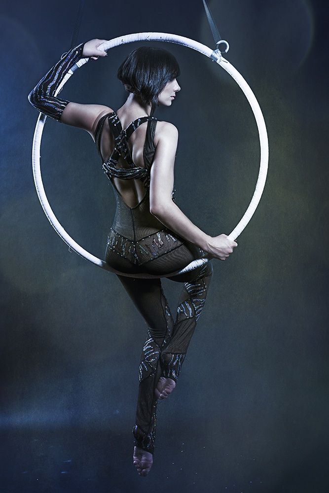 a woman in a black outfit with silver detailing is posing in an aerial hoop. she is holding onto the hoop with her left hand and her right hand is extended out to the side. the background is dark blue. she looks alluring and graceful.