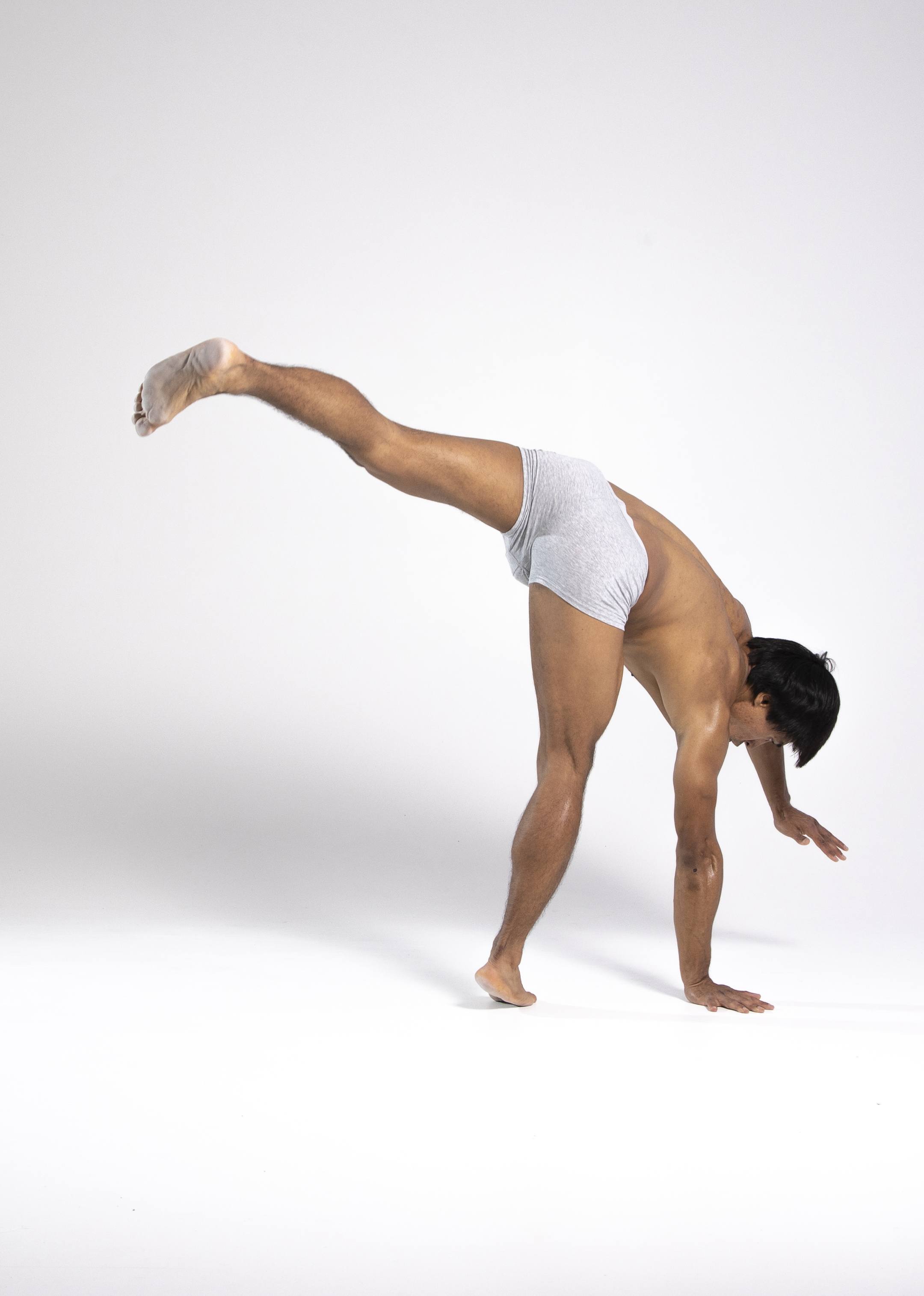 a shirtless man is performing a yoga pose indoors against a white background. he is wearing light grey underwear and is barefoot. his body is bent forward with his hands on the ground while one leg is extended upwards to maintain balance. he appears to be engaged in a fitness routine that requires flexibility and strength.