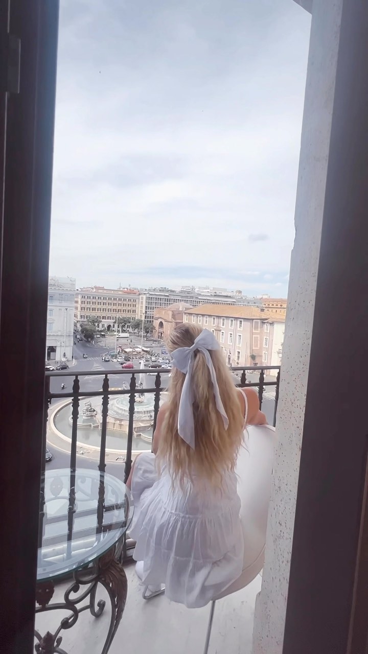 a woman with blonde hair and a white hair bow sits on a white chair on a balcony, looking out at a city view. she is wearing a white dress. in the distance, there are buildings, a fountain, and traffic. the balcony has a decorative iron railing.