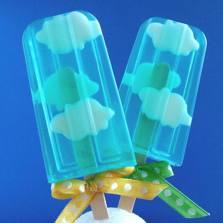 two blue popsicles with white clouds and yellow and green ribbons tied around the sticks. the popsicles are set against a bright blue background. they appear to be homemade.