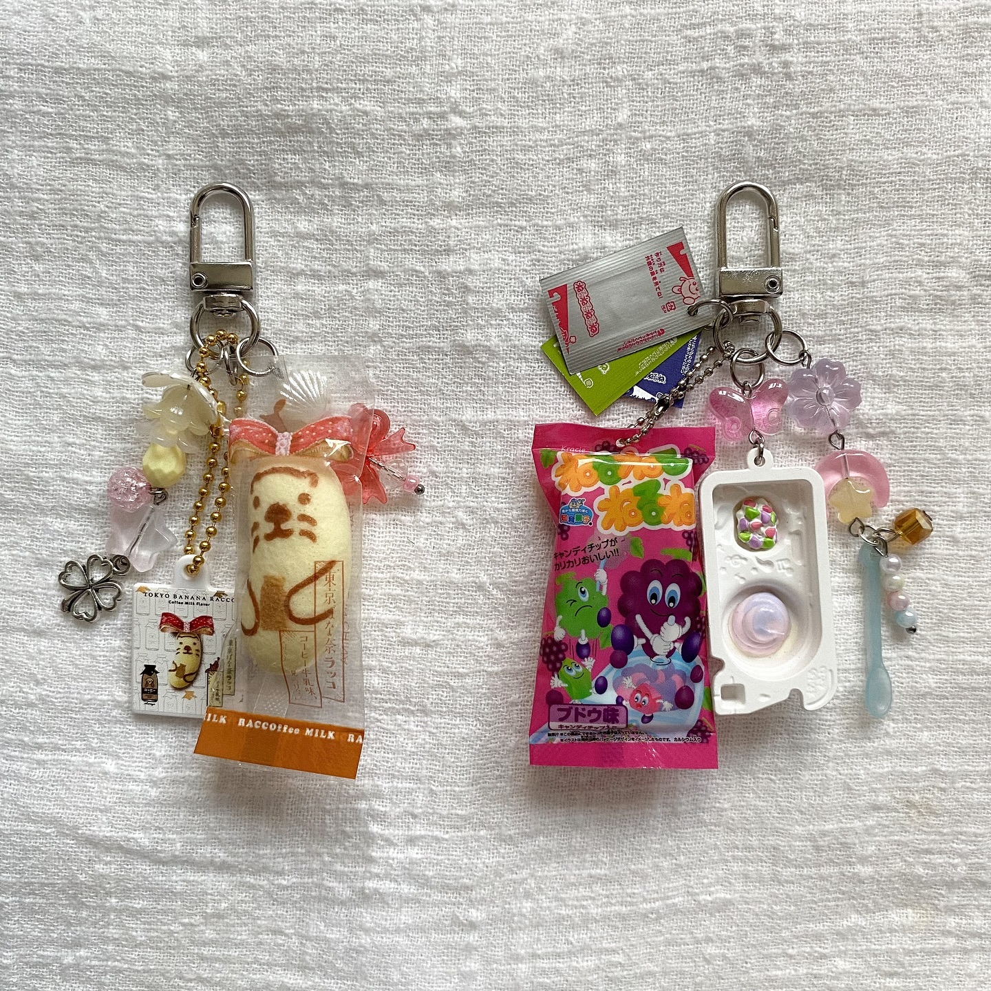 the image shows two keychains on a white textured surface. the keychain on the left features a miniature tokyo banana treat, a small box with an image of a cartoon raccoon, beads, and a fourleaf clover charm. the keychain on the right features a pink grapeflavored candy package, a small plastic mold, beads, a miniature spoon, and a few small green and white cards or packets. the keychains are both attached to silver clasps.
