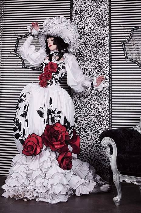 a woman wearing a beautiful white and black gothic dress with ruffles, a large red rose and lace trim. she is posing in front of a striped background with a black and white patterned wall. the dress is accessorized with red roses, and she is wearing a large white lace hat. she is standing next to a vintage chair with a black cushion.