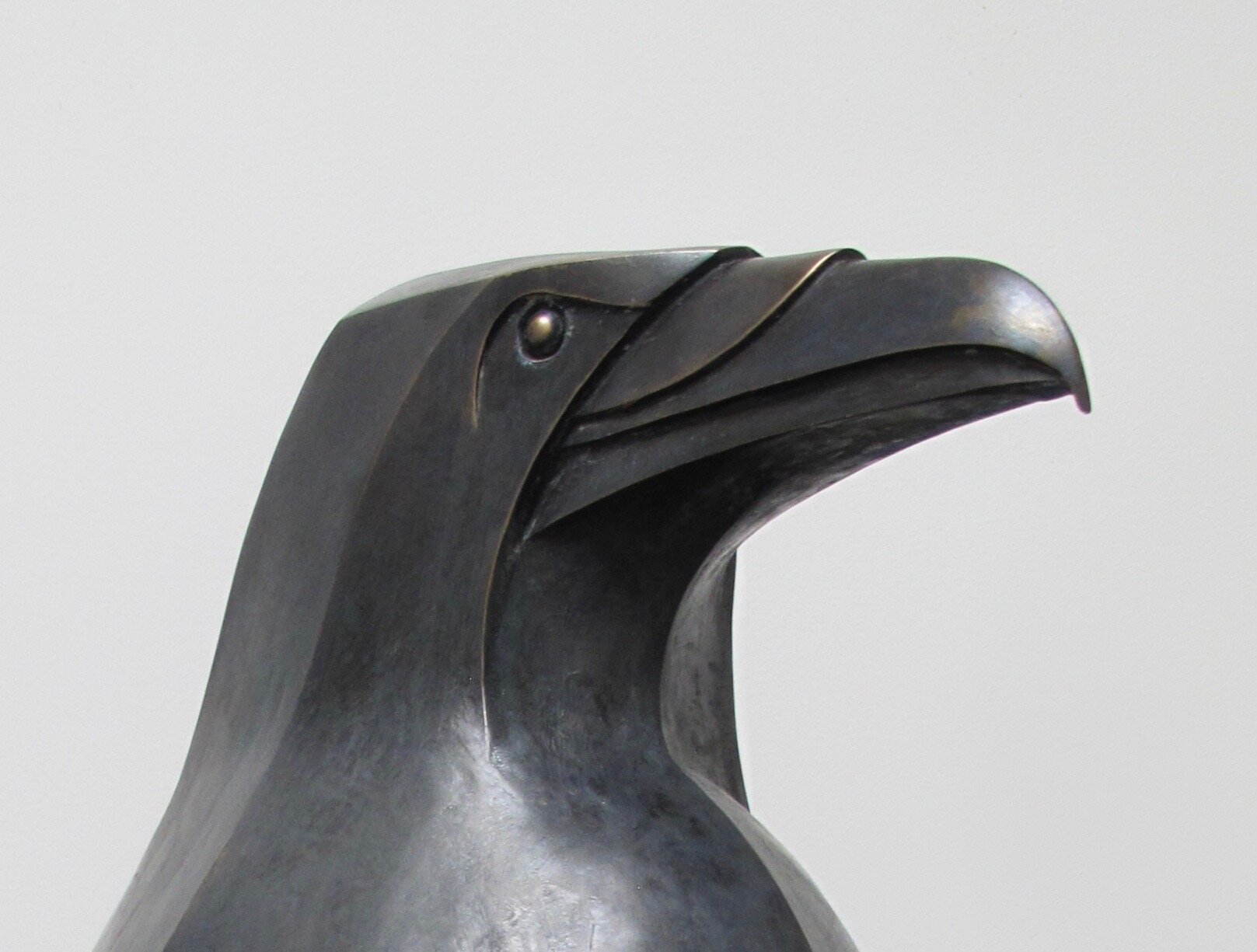 a bronze sculpture of a bird's head, resembling a raven. the sculpture is modern and abstract, with a metallic finish. the bird is shown in profile, highlighting the shape of its head, beak, and eye. the sculpture has a dark, almost black color.