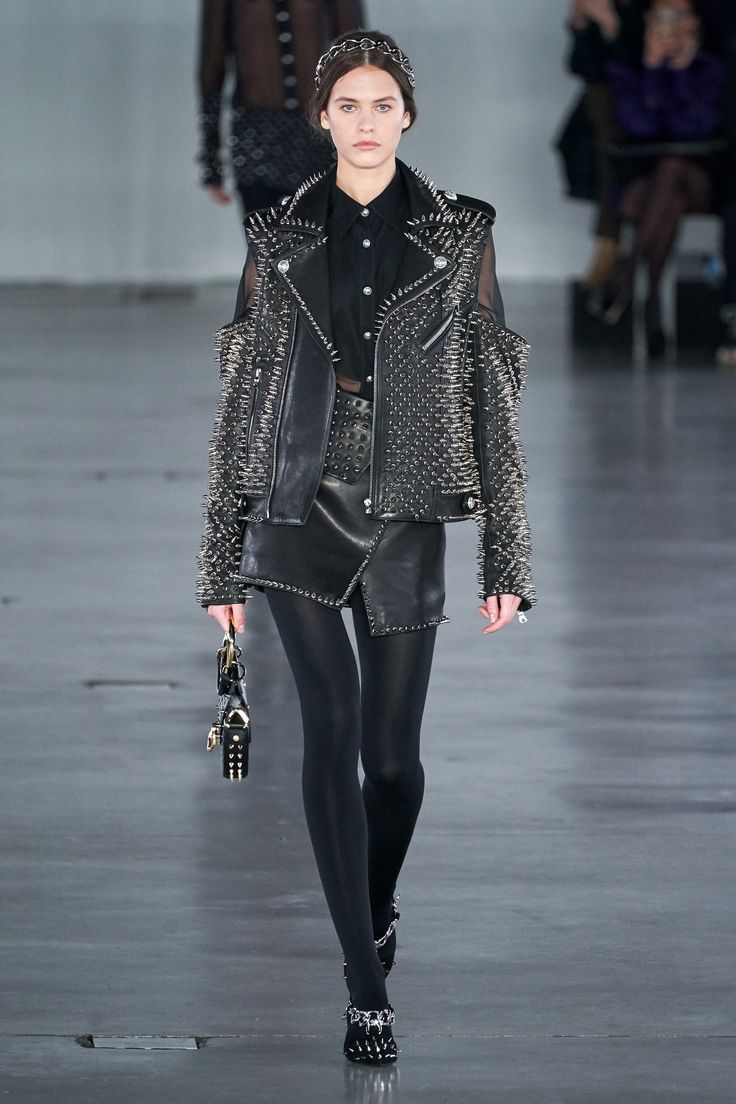 a full shot of a female model walking down a runway. she is wearing a black studded leather jacket, a black leather mini skirt, black tights, and black shoes with embellishments. she also has on a black shirt and carries a small handbag. the model's hair is styled back, and she is wearing a thin headband. the background is blurry, but shows other people watching. the overall aesthetic is punk or rockinspired.