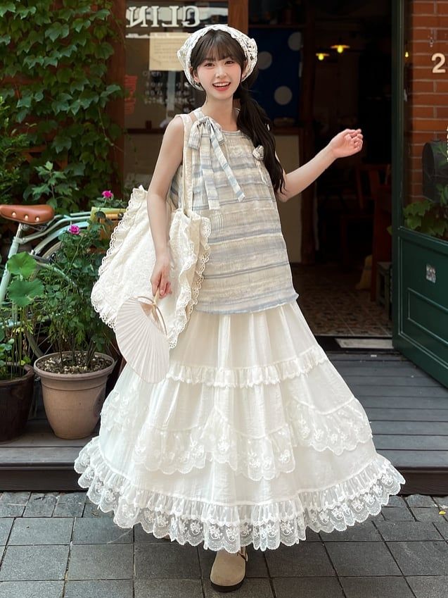 a woman wearing a blue and white striped top, a flowy white lace skirt, and a white crochet hat is posing outdoors. she is holding a white crochet bag and a folding fan. the background shows a street scene with a building and plants. the image is brightly lit with natural light.