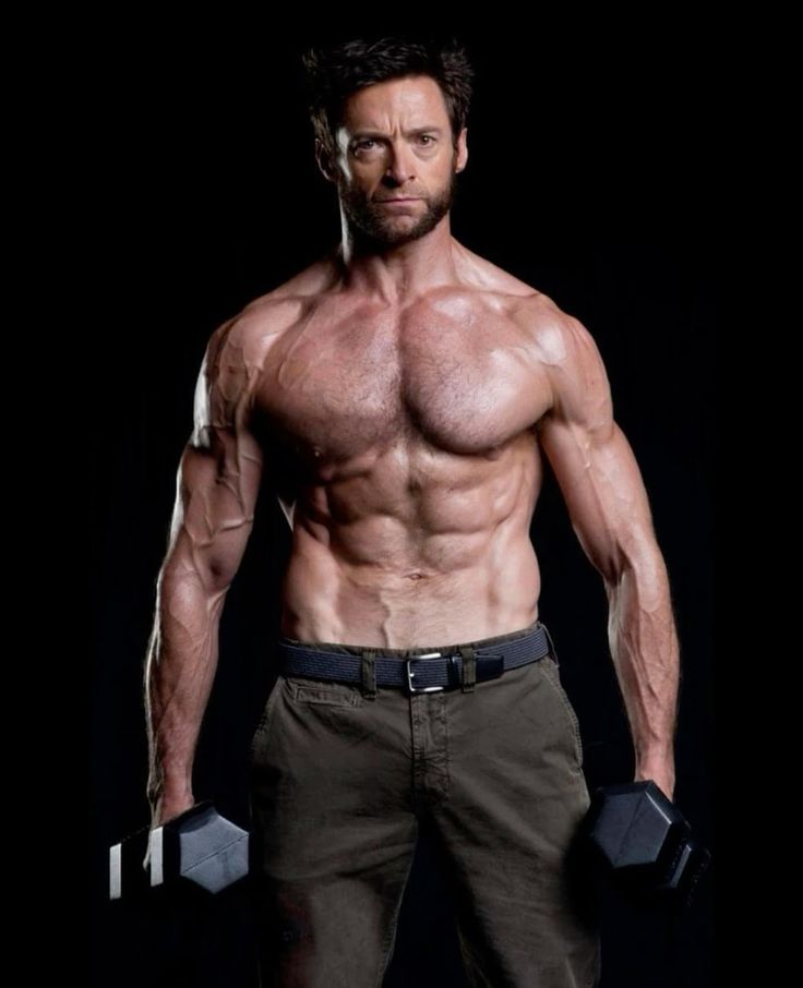 a shirtless man with a muscular physique holds a dumbbell in each hand. he is looking directly at the camera with a serious expression. he has a sixpack and is very fit. he is posing in a studio setting. the man's name is hugh jackman and he is known for his role as wolverine in the xmen film franchise.