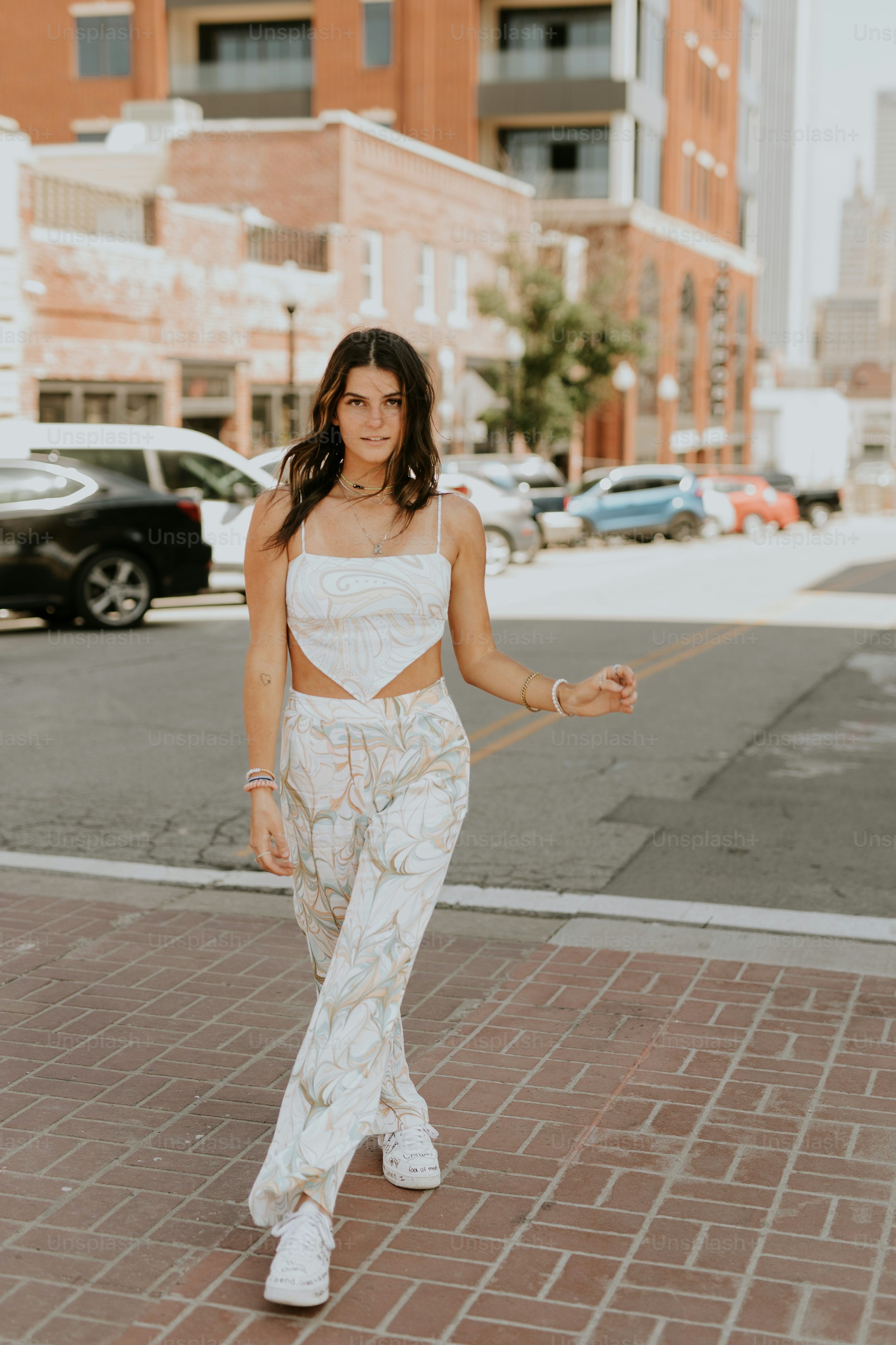 a young woman strolls down a city street, confidently showcasing her stylish summer outfit. she's wearing a white floral print pantsuit with a cropped top, white sneakers, and delicate jewelry. her long brown hair flows naturally, and her overall demeanor is one of relaxed confidence. the urban background features brick buildings and parked cars, adding context to the scene. the bright lighting and vibrant colors enhance the overall aesthetic.