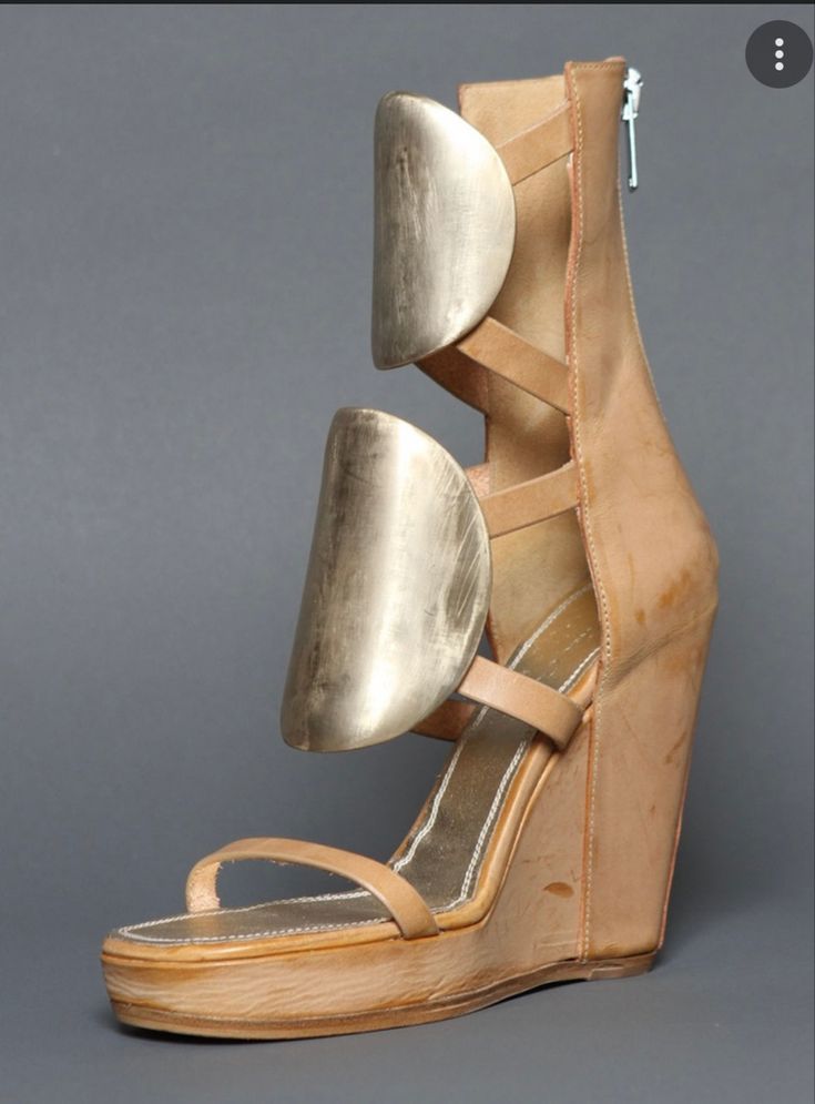 a closeup shot of a pair of women's beige leather wedge sandals. the sandals feature unique gold metallic accents that are semicircular in shape and attached to the straps.  the sandals have ankle straps and open toes, and sit on a substantial wedge heel. the overall style is modern and sophisticated.  the background is a neutral gray, providing contrast to the shoe's color and texture.