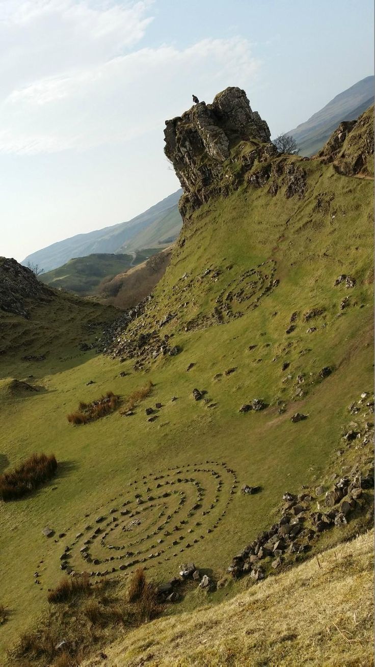 the image shows a mountainous landscape with a green valley. there is a rock formation at the top of a hill. in the valley, there is a spiral made of rocks. a bird is perched on top of the rock formation.