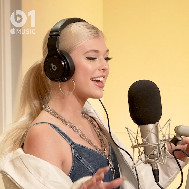 a woman with blonde hair is singing into a microphone while wearing headphones in a studio setting. she is also wearing a denim top, a white jacket, and jewelry, including a chain necklace and hoop earrings. there is an apple music logo in the top left corner of the image.