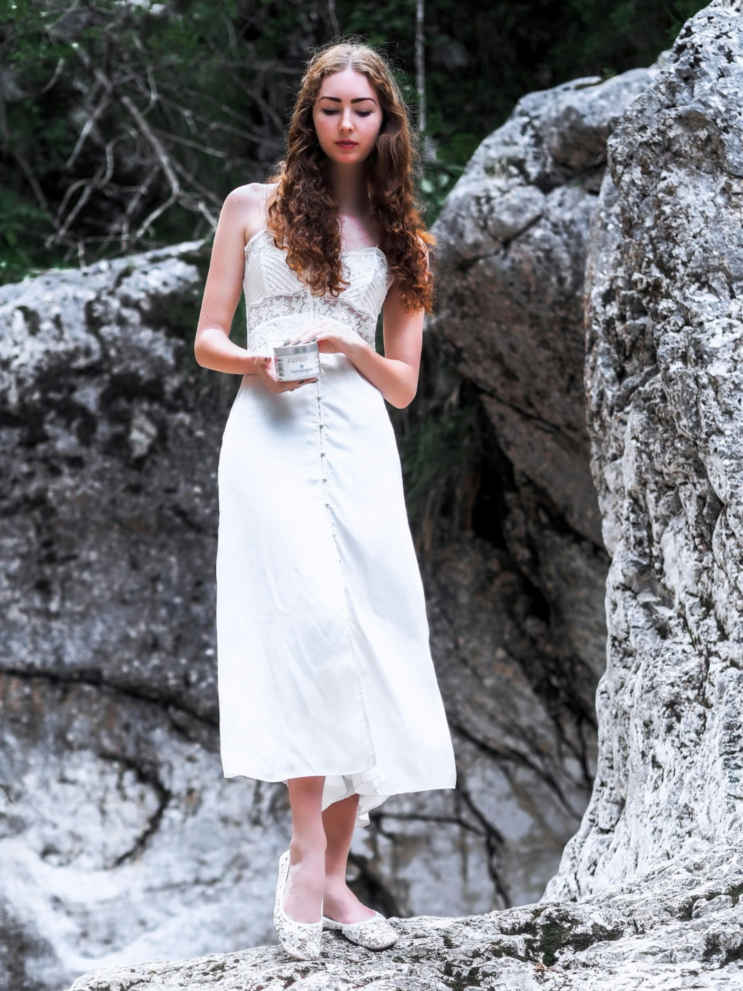 a young woman with long curly brown hair is standing outdoors among rocks and trees. she is wearing a white lace maxi dress and white flats. she is holding a small white box. the overall mood is serene and peaceful. the image has a mystical or fairytalelike quality.