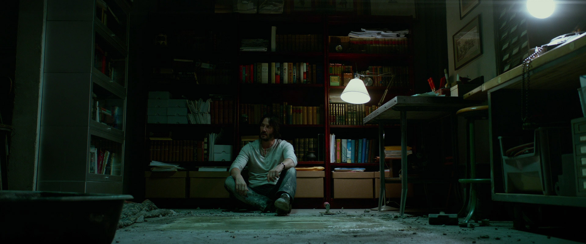 a man sits on the concrete floor in a dark and mysterious room, surrounded by bookshelves and a table with a lamp on it. he is alone and appears to be lost in thought. the room is dimly lit, creating a sense of mystery and intrigue. this image appears to be a still from a film.