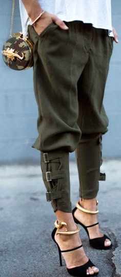 a woman wearing green cargo pants and black high heels with gold ankle bracelets. she is holding a camouflageprint purse. she is standing on a street, looking chic and trendy.