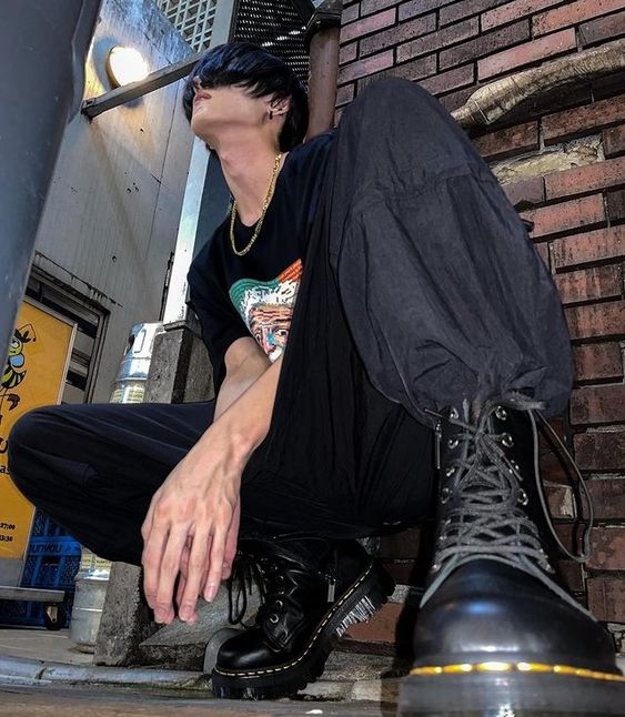 a young man with black hair, wearing a black tshirt and black pants, sits in an alleyway with brick walls. he wears black dr. martens boots and looks up towards the light source. the overall aesthetic leans towards grunge fashion. the photo is taken from a low angle, emphasizing the height of the subject and the towering brick walls.