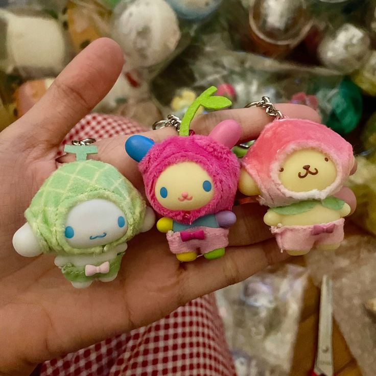 a hand holds three sanrio character plushie keychains. the characters are cinnamoroll (green), my melody (pink), and pom pom purin (peach). they are all wearing costumes and attached to keychains.