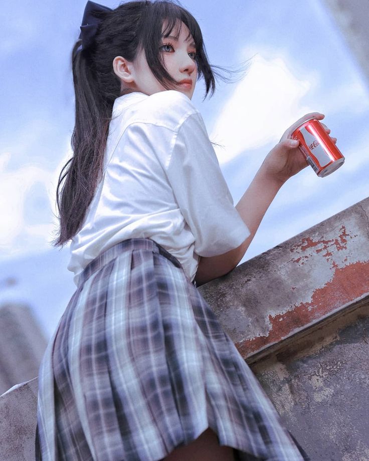 a young asian woman with long black hair tied in a ponytail, wearing a white shirt and a gray and white plaid pleated skirt, holds a can of cocacola while standing against a weathered concrete wall under a partly cloudy sky. she is looking away from the camera, her expression is calm and thoughtful.  the overall tone of the image is soft and dreamy.