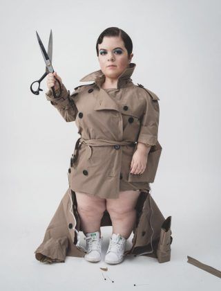 the image shows a stylish young woman with short stature, possibly due to dwarfism or achondroplasia, modeling a beige trench coat. she is holding a pair of large scissors, adding a dramatic element to the pose. the woman is wearing white sneakers and the trench coat is oversized and stylishly styled. the background is plain white, which helps the model and clothing stand out. the overall style evokes highfashion editorial photography.