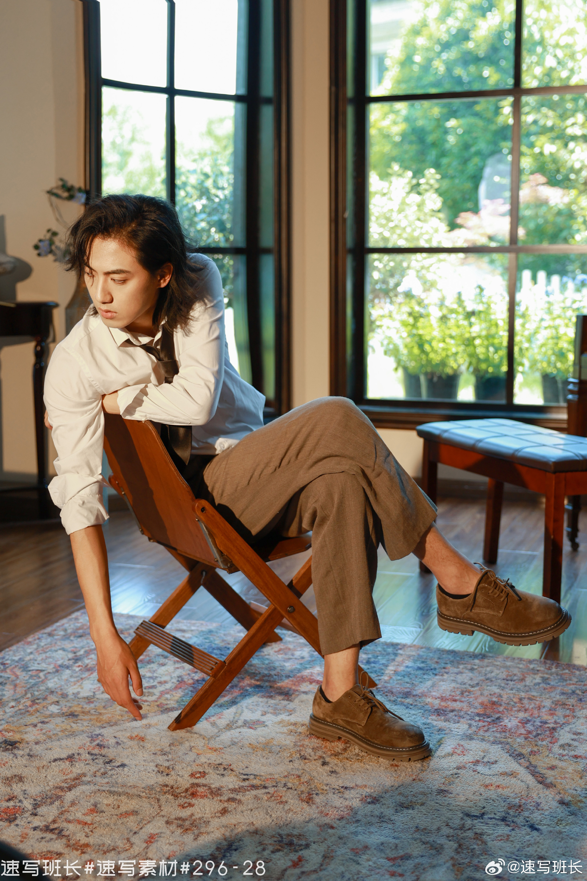 a young man with long hair sits casually on a wooden folding chair, his left arm draped over the back of the chair, his right hand resting on the floor. he is wearing a white buttondown shirt, brown trousers, and brown shoes. the room is sunlit, with a large window in the background and a patterned rug on the floor.