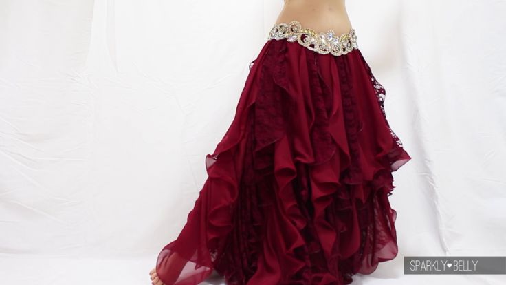 a woman is shown from the waist down, wearing a red chiffon belly dance skirt with multiple layers of ruffles. the skirt is held up by an ornate belt adorned with jewelry and sparkles. the backdrop is a plain white sheet.