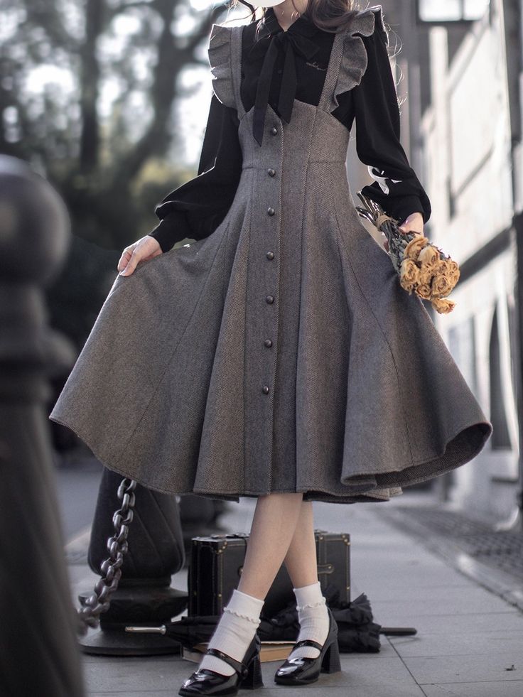 a woman in a stylish gray dress and black blouse poses on a city sidewalk. the dress has a long, flowing skirt, buttondown front, and ruffle details on the shoulders. the woman is also wearing black shoes and white socks. she is holding a bouquet of yellow roses in her left hand. the background of the image shows a city street with buildings and a sidewalk. the lighting is natural and casts shadows on the ground. the overall tone of the image is stylish and elegant.