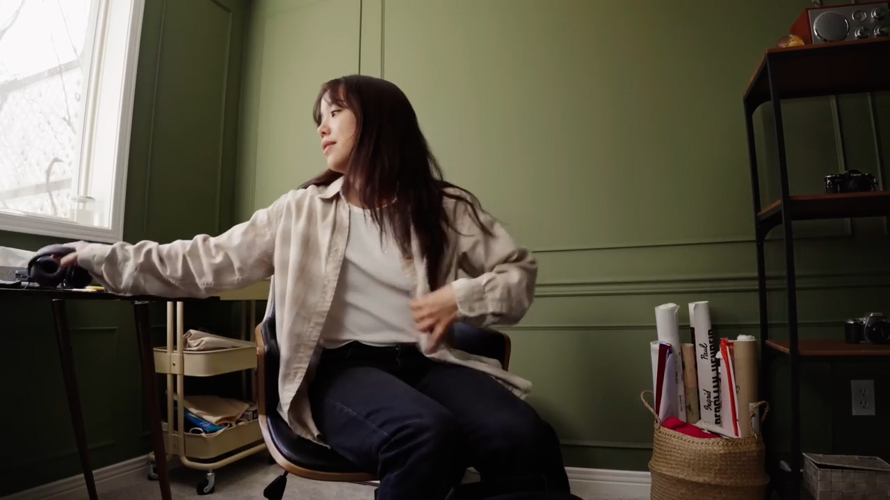 a young woman is shown in her home office, seated in a black chair.  she is in the process of putting on or adjusting a light beige overshirt. the room is bright, lit by a window to the left, and decorated in a minimalist style with muted green walls.  a desk with organizational elements is visible, as well as a bookshelf.