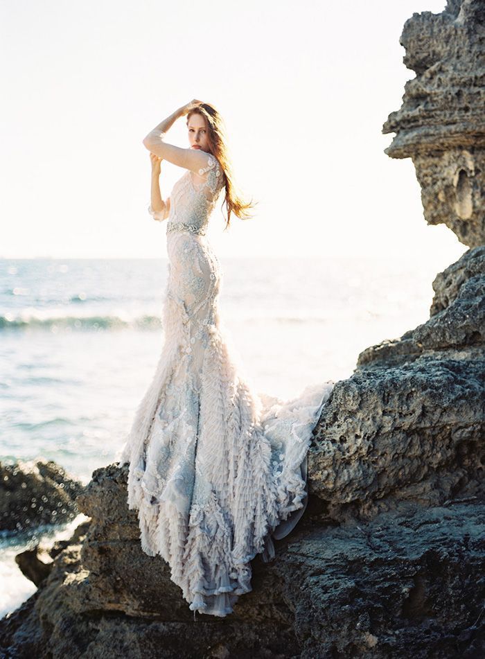 a bride stands on a cliff overlooking the ocean. she is wearing a beautiful white gown with a long flowing skirt. her hair is flowing in the breeze and she looks out over the water. the sun is shining and the sky is a bright blue. the scene is romantic and picturesque.