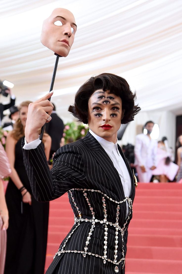 ezra miller attends the met gala, wearing a pinstripe suit with a jeweled corset, theatrical multieyed makeup, and holding a mask of his own face. he's on the red carpet, striking a pose.