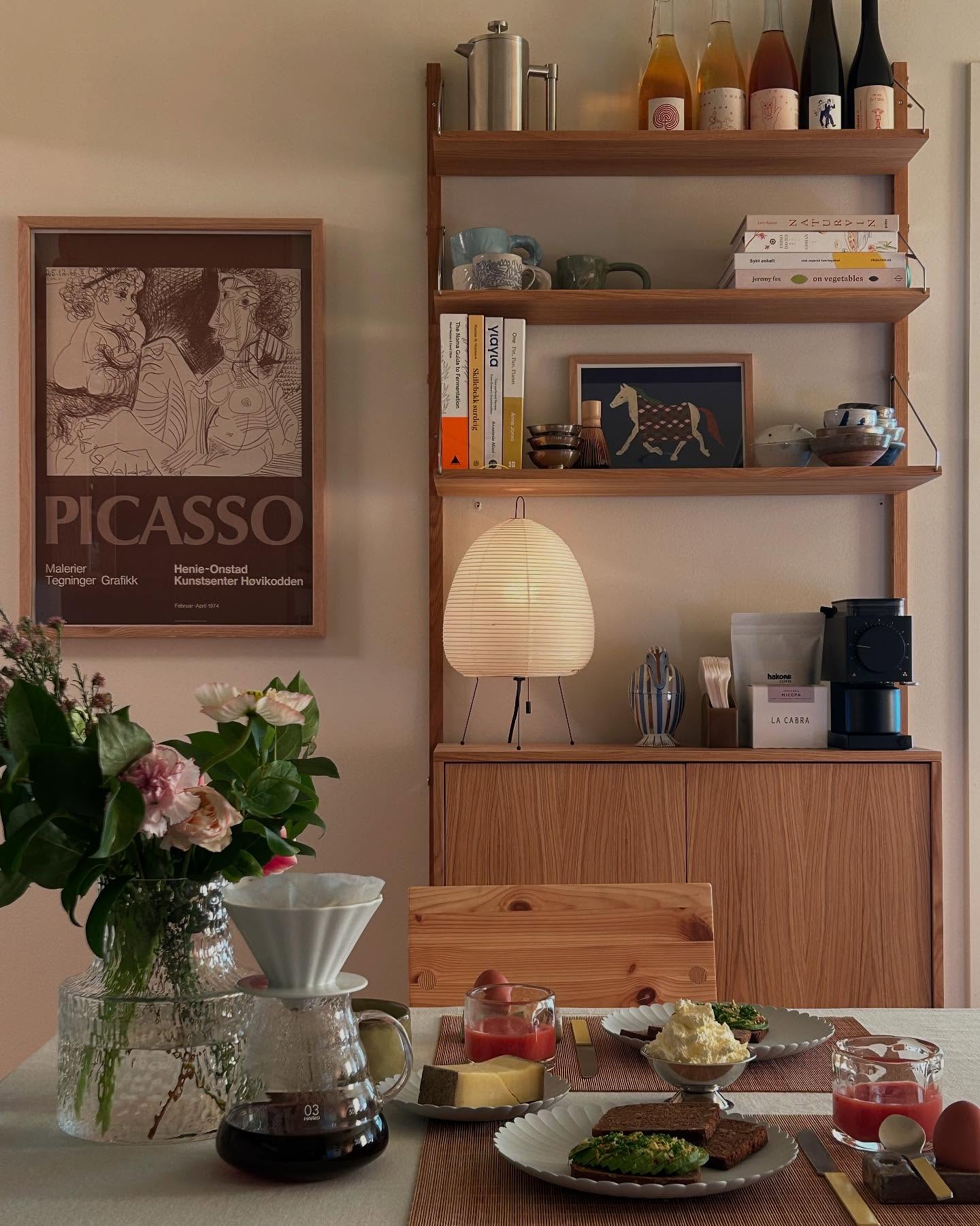 a shot of a beautifully arranged breakfast table in a welllit room. a vase filled with flowers sits beside a pour over coffee setup. two place settings are visible, each with a plate of food, a glass of red liquid, and utensils. a picasso poster hangs on the wall next to a shelving unit filled with books, cups, and various items. a modern lamp illuminates the scene, and a wooden cabinet is below the shelves, holding a coffee grinder and a bag of la cabra coffee. a chair is partially visible behind the table.