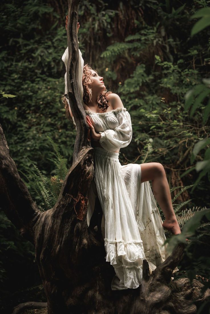 a woman in a long, flowing white offshoulder dress sits on a large tree branch in a lush forest. she has curly hair and is looking up with a dreamy expression. her bare foot is visible, and the surrounding forest is dense with ferns and other greenery. the overall mood is ethereal and romantic.