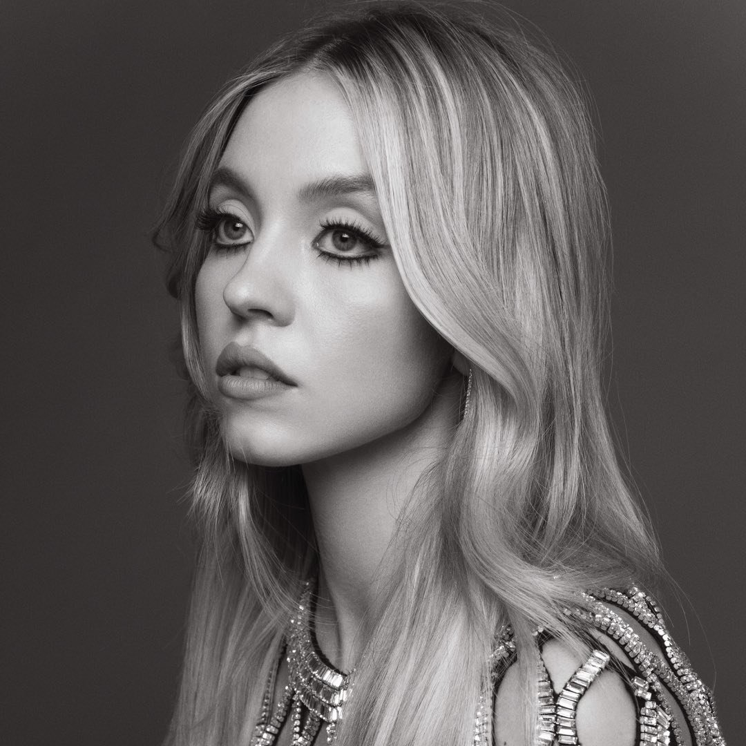 a black and white portrait of sydney sweeney with blonde hair, wearing jewelry and heavy eye makeup, looking to the side.