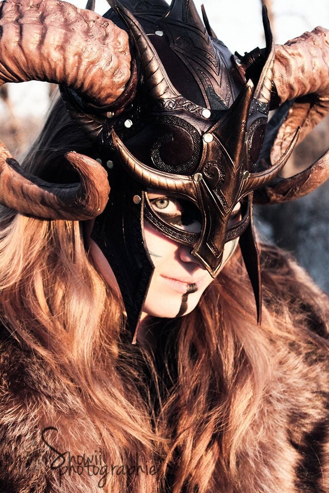 a person in fantasy cosplay attire featuring a horned helmet and fur clothing. the helmet is dark with intricate designs and large, curved horns. the person has makeup applied, including dark markings on their face. they are wearing fur clothing, and the overall aesthetic is reminiscent of a viking or nordic warrior from fantasy games like the elder scrolls: skyrim.
