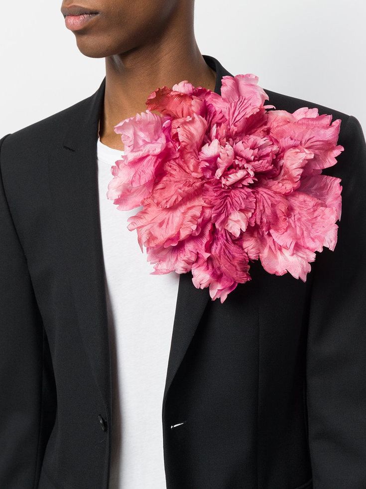 closeup shot of a male model wearing a black blazer with a large, pink fabric flower brooch pinned to the lapel. the flower is a prominent feature, appearing to be a stylized peony with many layers of petals. the model is wearing a white undershirt, and the focus is sharply on the flower and its placement on the jacket.  the image showcases highfashion details and styling.