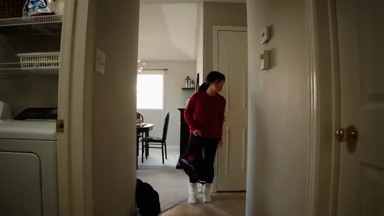 a young woman is entering her home, carrying a black bag. she is wearing a red sweater, black pants, and white boots. the image is taken from the perspective of someone inside the house, looking towards the front door. the hallway is narrow and leads to the laundry room on the left and the rest of the house on the right. the lighting is natural, coming from the window in the background.