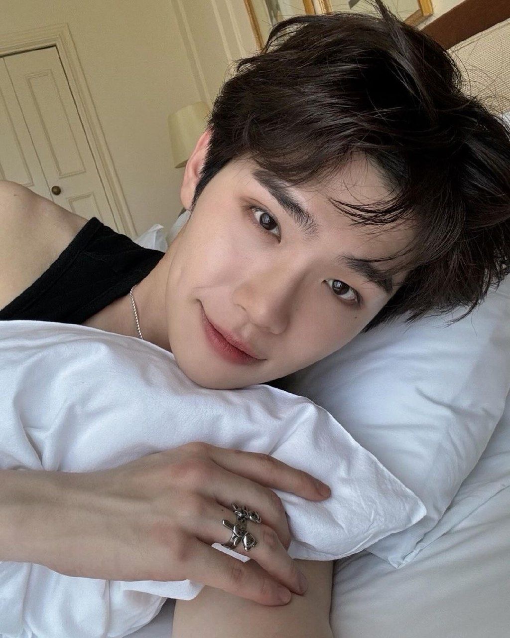 the image shows a young man with dark hair lying on a bed, his head resting on a white pillow. he's wearing a black tank top and has a slight smile on his face. the man is looking directly at the camera, creating a sense of connection with the viewer. the lighting is soft and natural, likely coming from a window, and highlights the man's features. he is wearing a large silver ring on his left hand. the overall feeling of the image is one of calmness and relaxation.