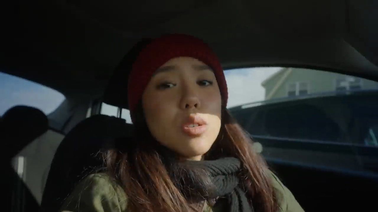 young asian woman with a red beanie and scarf is taking a selfie video inside a car. the sun is shining brightly, illuminating her face. the car appears to be parked on a residential street.