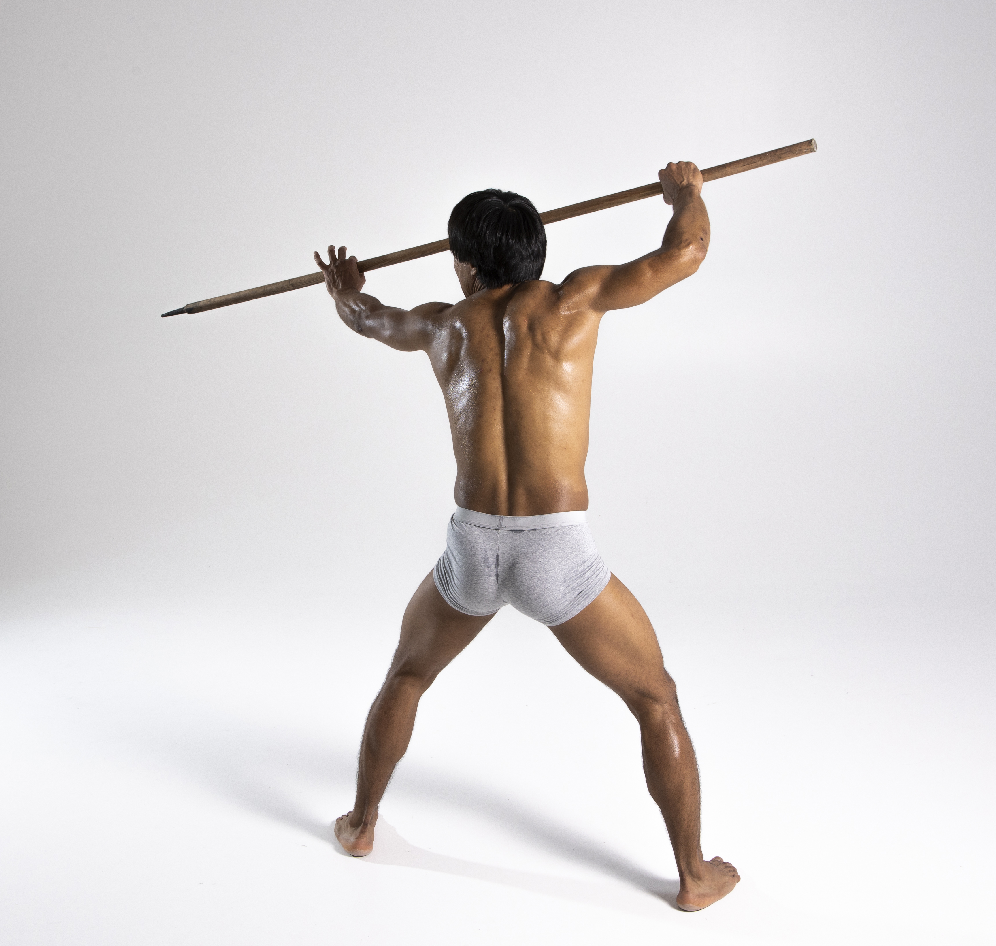 a barechested man standing with a spear held over his head, facing away from the viewer. he is wearing gray underwear. the background is a plain white studio backdrop. his body appears muscular and athletic.