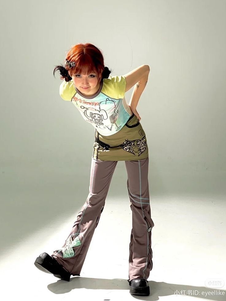 a young woman with red hair stands in a studio with a white background. she's wearing a light green tshirt with a cartoon design, brown baggy pants, and black boots. she has a cute expression on her face and is looking at the camera. she's posing casually, slouching her shoulders a little bit. she's wearing her hair long with bangs.