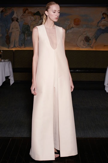 a woman in a long white dress poses elegantly in a studio setting. her dress is simple and sleek, highlighting the clean lines and sophisticated design. the model's poised stance and confident gaze convey a sense of formal elegance. the white backdrop emphasizes the stark beauty of the dress and the model's natural features.