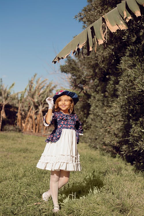 a young girl, perhaps 68 years old, stands in a grassy field, radiating joy.  she wears a charming white dress with a blue floral bodice and several layers of ruffles.  the dress is reminiscent of vintage or victorian styles.  a matching blue hat with floral accents adorns her head, which she delicately holds with a white glove. her expression is sunny and playful. the background features lush green grass and vegetation under a clear blue sky, with the remnants of a dried banana leaf adding a slightly exotic touch to the scene.  the overall mood is light, airy, and cheerful.