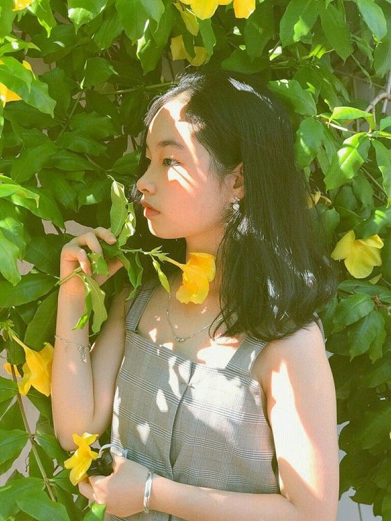 a young woman with shoulderlength black hair is standing in front of a wall of green leaves and yellow flowers. she is wearing a gray plaid sleeveless top and has a yellow flower tucked behind her ear. she is holding a stem of leaves in one hand and a yellow flower in the other. the sun is shining on her face, creating a dappled light effect.