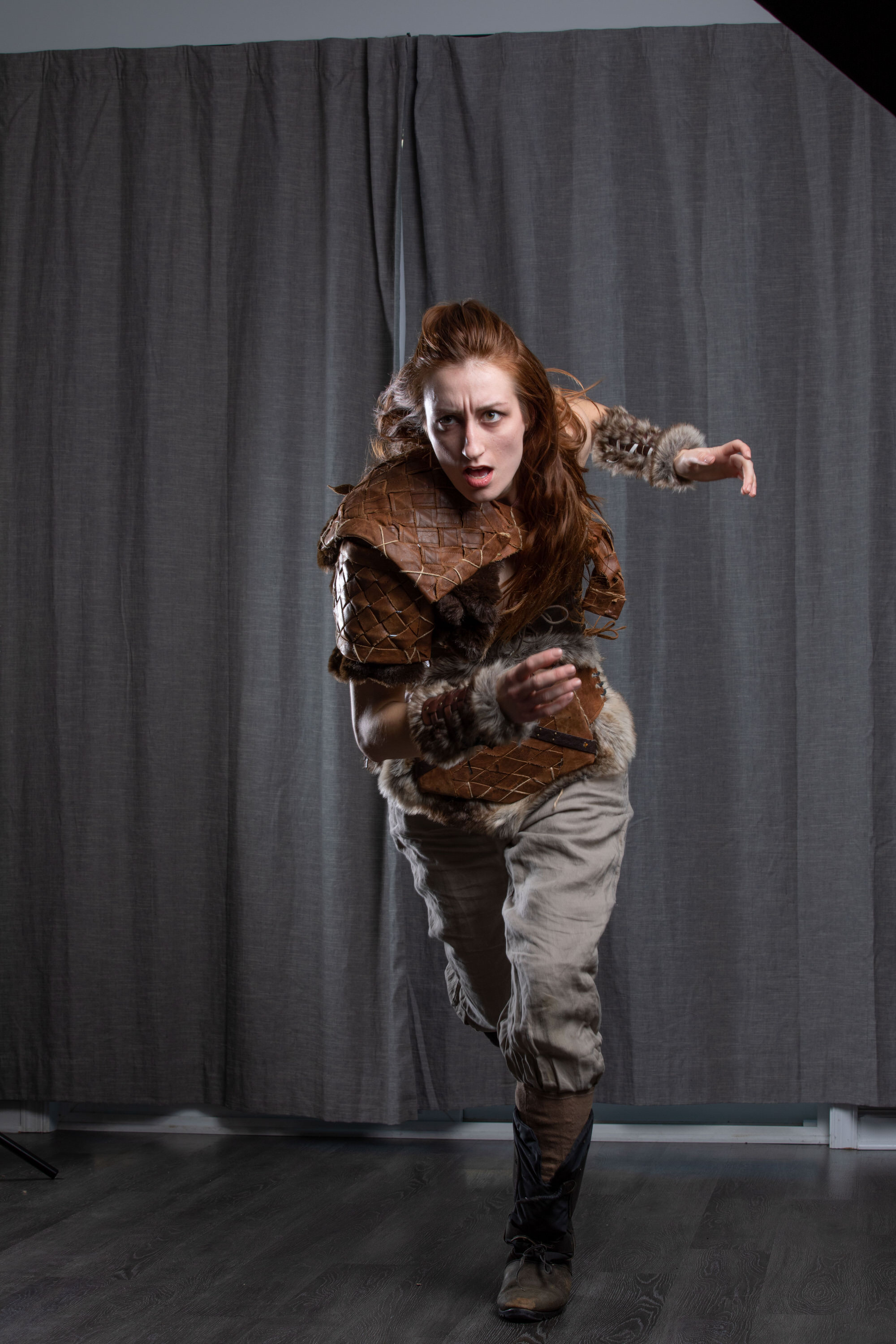 a woman with long red hair is captured midrun against a backdrop of grey curtains. she is dressed in a warrior costume, featuring a brown leather breastplate and arm guards, suggesting a fantasy or historical character. her expression is one of intense focus and determination. the lighting is artificial, casting subtle shadows and highlighting the textures of her costume.